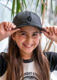 A young girl wearing a black baseball cap with the UJA flame logo engraved in black.