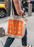 A woman walking in NYC with a beige tote bag with an orange UJA square logo.