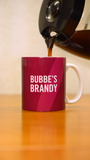 A coffee pot pouring coffee into a pink mug that says "Bubbe's Brandy" in white letters.