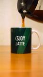 A coffee pot pouring coffee into a green mug that says "(S)oy Latte)" in white letters.