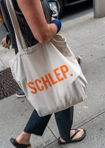 A woman in NYC walking with a beige tote bag that says "Schlep" in orange letters. 