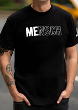A man on the streets of NYC wearing a black shirt that says "Mensch" with the letters "Me" bolded. 
