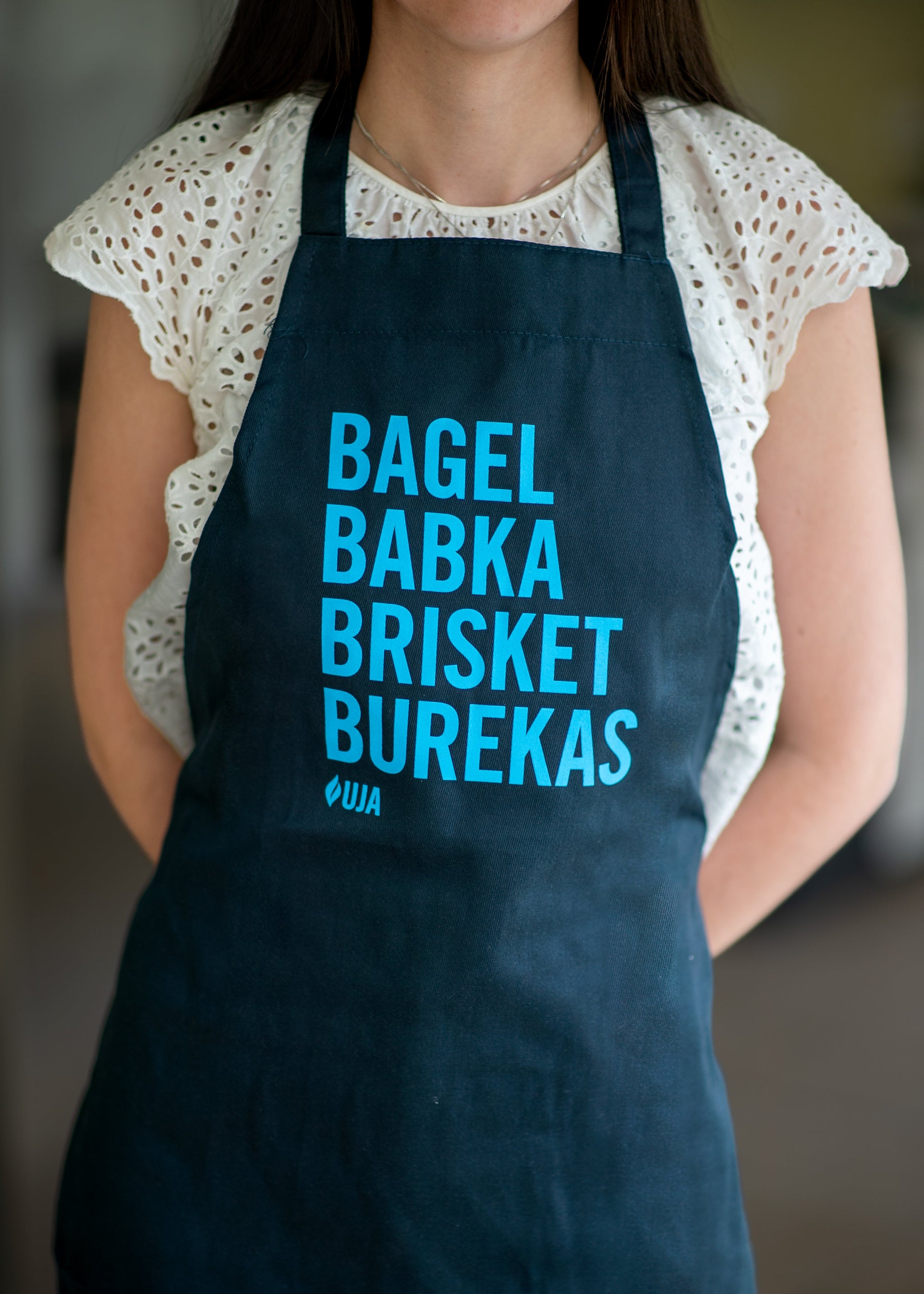 Woman wearing a black apron that says "Bagel, Babka, Brisket, Burekas" in bright blue letters. 