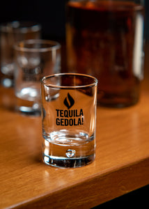 Tequila Gedola shot glass on a wooden surface with other glasses in the background.