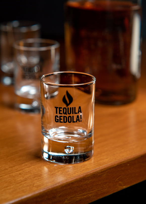 Tequila Gedola shot glass on a wooden surface with other glasses in the background.