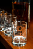 Row of Tequila Gedola shot glasses on a wooden surface with a bottle in the background.