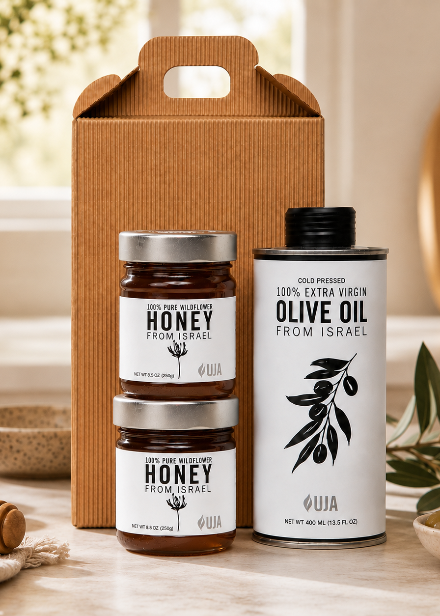 A gift set featuring two jars of UJA's honey and one bottle of olive oil, arranged in front of a brown cardboard gift box on a light stone surface with olives, a honey dipper, and greenery in a softly lit kitchen setting.