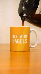 A coffee pot pouring coffee into a yellow mug that says "Best with Bagels" in white letters. 