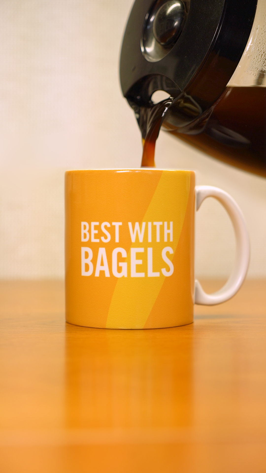 A coffee pot pouring coffee into a yellow mug that says "Best with Bagels" in white letters. 