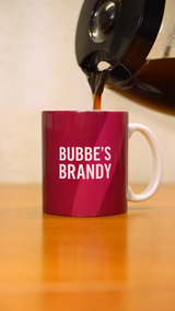 A coffee pot pouring coffee into a pink mug that says "Bubbe's Brandy" in white letters. 