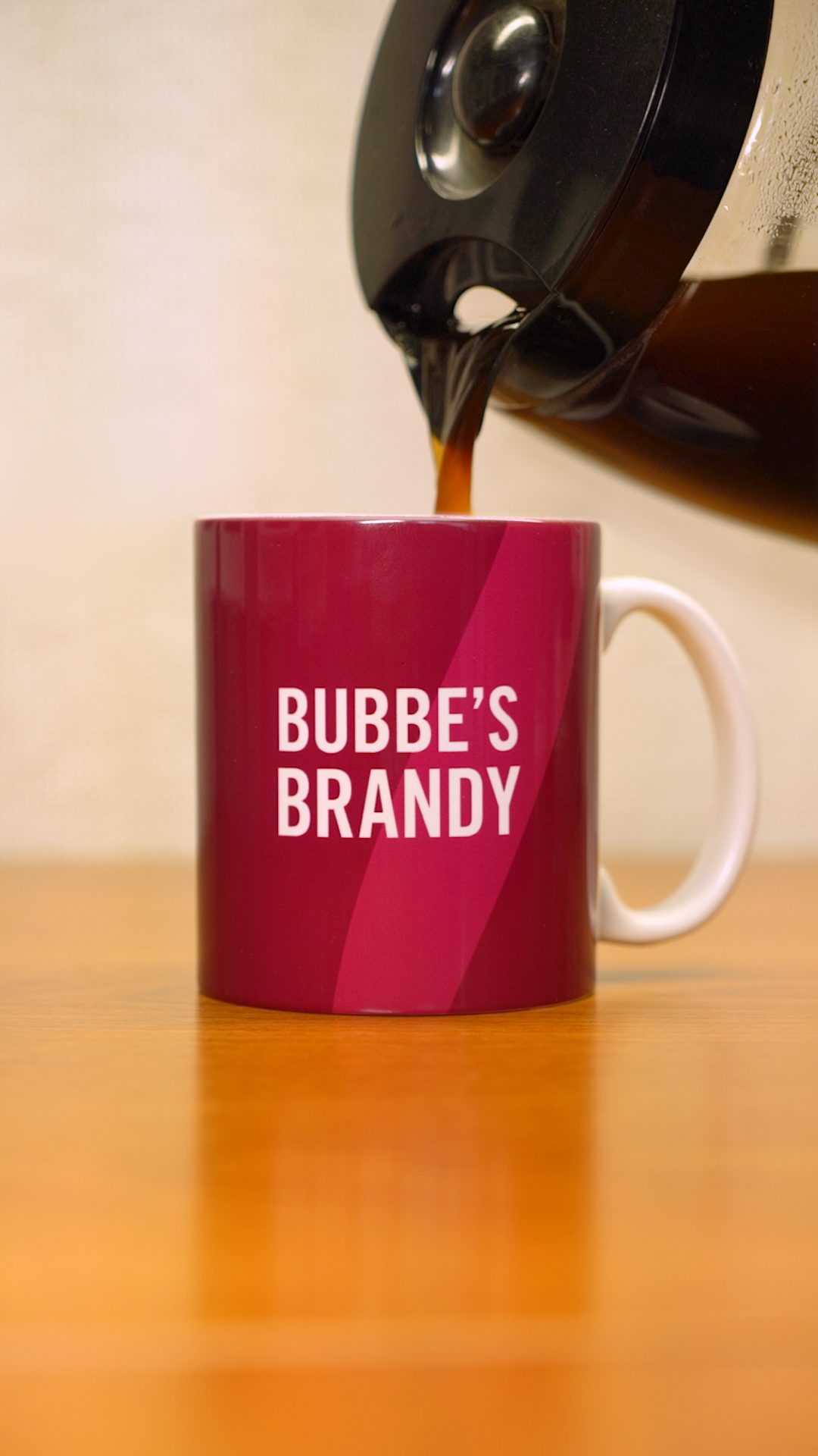 A coffee pot pouring coffee into a pink mug that says "Bubbe's Brandy" in white letters. 