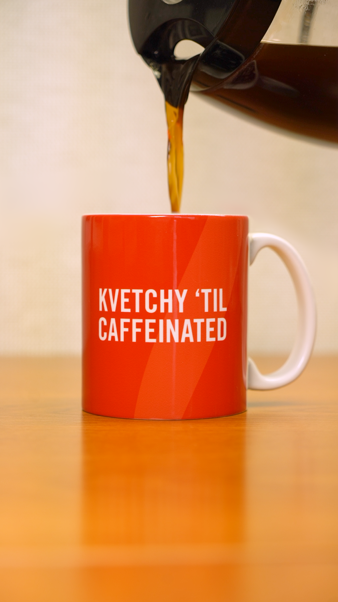 A coffee pot pouring into a burnt orange mug that says "Kvetchy 'Til Caffeinated" in white wording. 