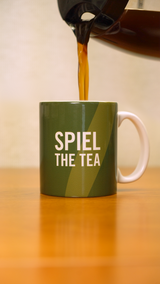 A coffee pot pouring coffee into a green mug that says "Spiel the Tea" in white letters. 