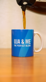 A coffee pot pouring coffee into a light blue mug that says "UJA & Me: The Perfect Blend" in white letters. 