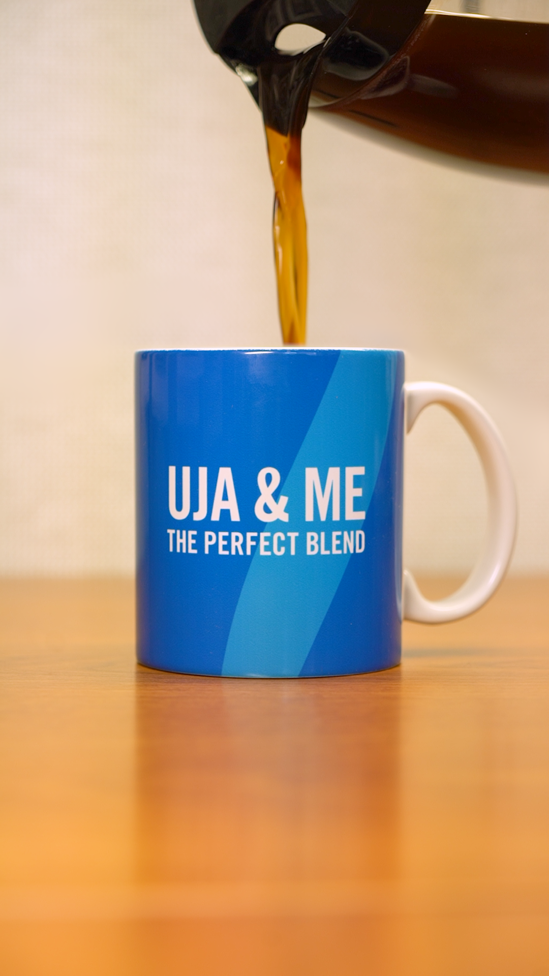A coffee pot pouring coffee into a light blue mug that says "UJA & Me: The Perfect Blend" in white letters. 