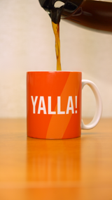 A coffee pot pouring coffee into a orange mug that says "YALLA!" in white letters. 