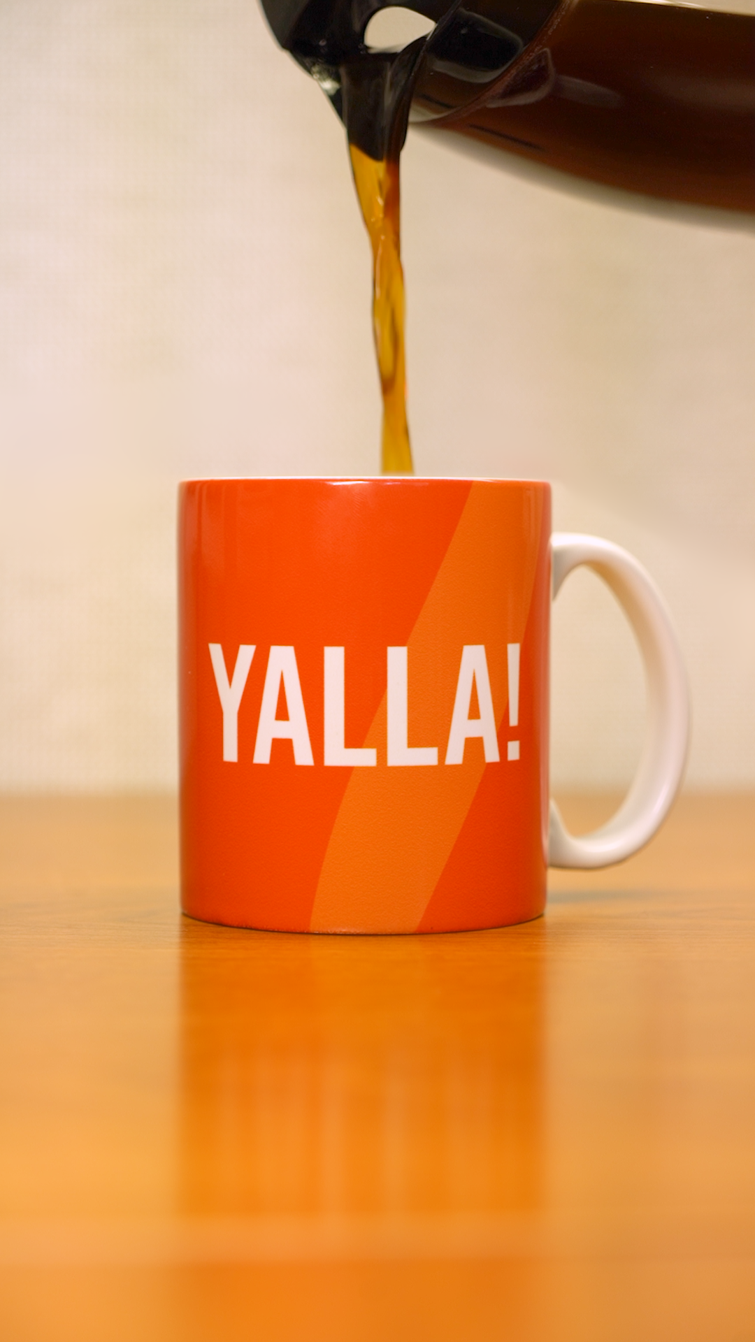 A coffee pot pouring coffee into a orange mug that says "YALLA!" in white letters. 